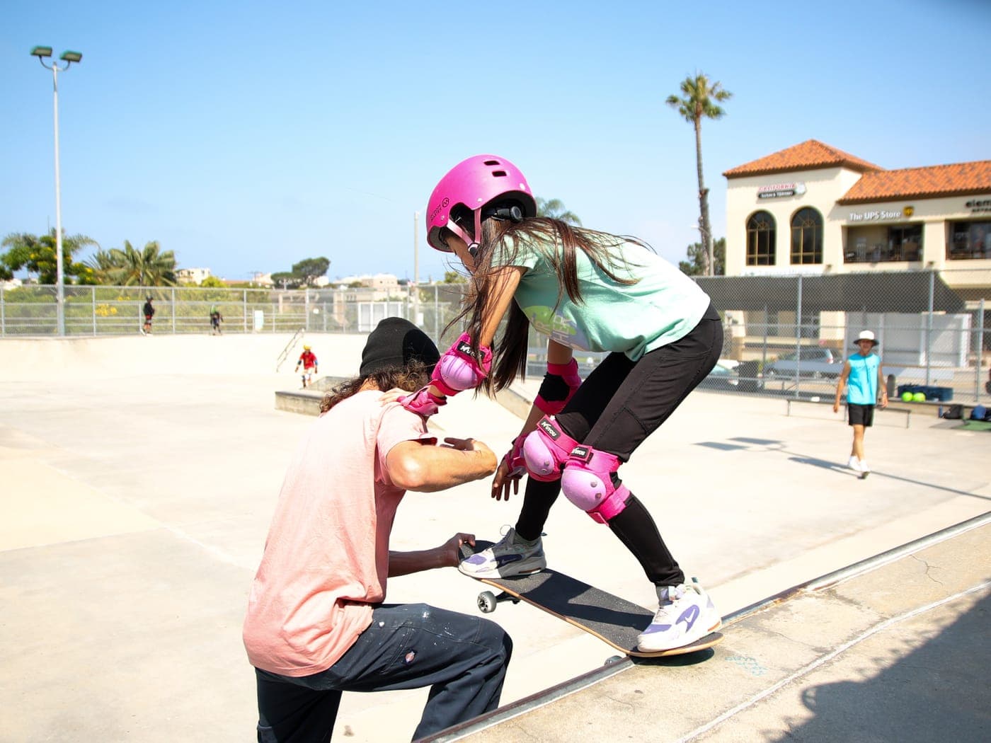 A BeachSports coach kneeling beside a young skater for one-on-one coaching at the Hermosa Beach Skatepark.