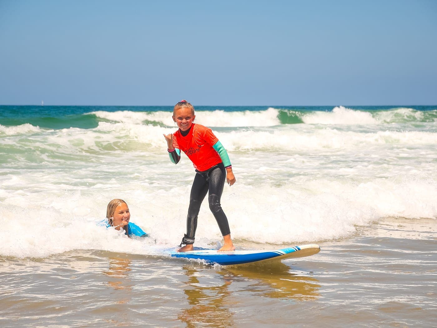 A young BeachSports camper riding a wave on a surfboard and throwing a shaka